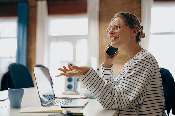 Smiling female entrepreneur working on laptop while sitting in office and talking by phone