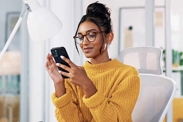 Office, Indian woman in glasses with smartphone for reading. Smile, businessperson on break with mobile, news application and chat on social media.