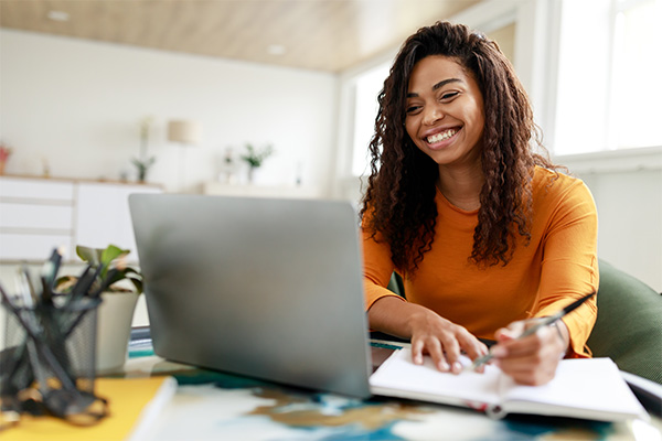 Smiling young woman sitting at a desk, using a laptop and writing notes in a notebook, representing focused study and academic effort.